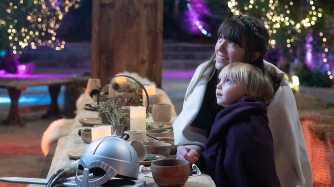 A woman and child wrapped in cloaks sit and look up towards the top left of the shot. They are at a long wooden table covered in wooden cups and bowls with a silver helmet in the foreground.